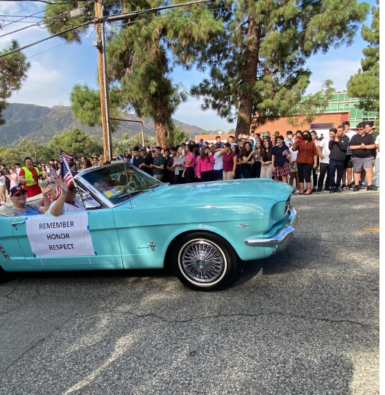 Turquoise car going by with REMEMBER, HONOR, RESPECT poster hanging, while the students, teachers, and Principal Kortoshian cheer them on. 

